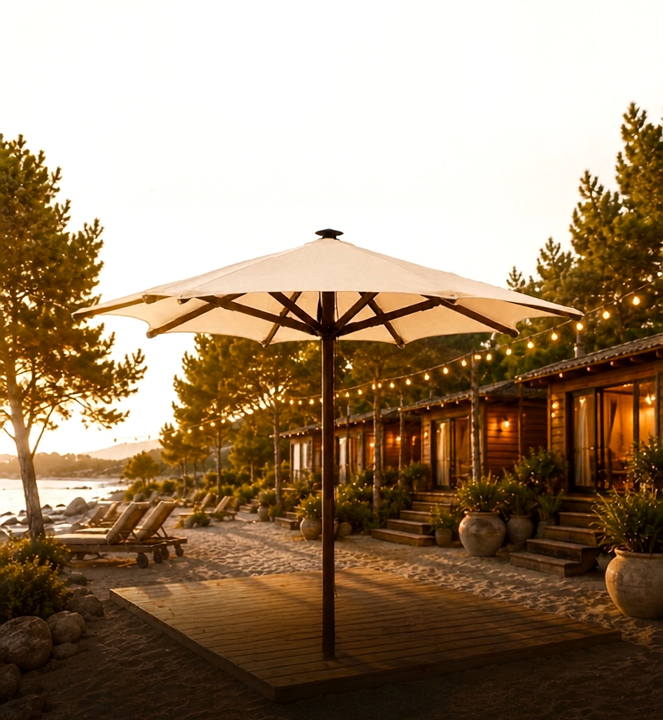 Parasol elegante de color crema sobre una plataforma de madera en una playa al atardecer, rodeado de tumbonas, cabañas iluminadas y árboles junto al mar.