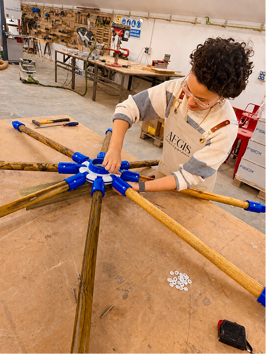 Artesana ensamblando las varillas de madera de un parasol AEGIS™ en el taller de fabricación, mostrando el proceso artesanal y el cuidado en cada detalle de la estructura.
